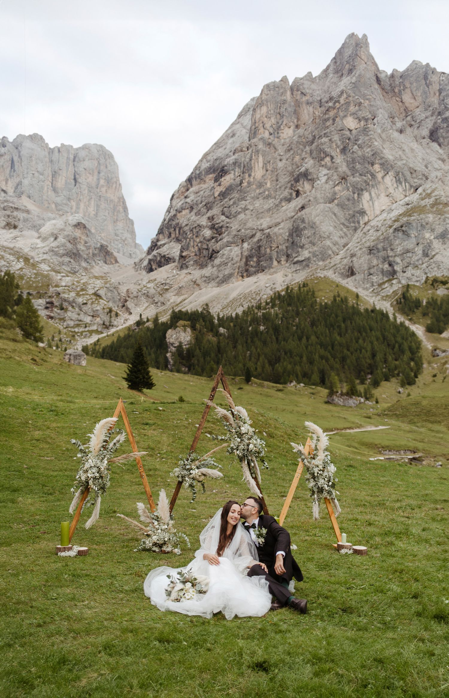 A couple poses on a grassy meadow with dramatic Dolomite mountain peaks rising in the background on their wedding day.