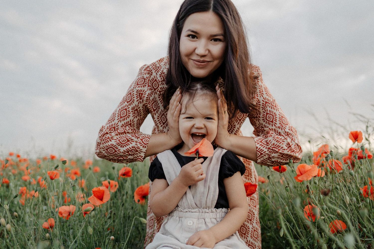 A parent and child share joyful moments together in a vibrant field of red poppies on a cloudy spring day.