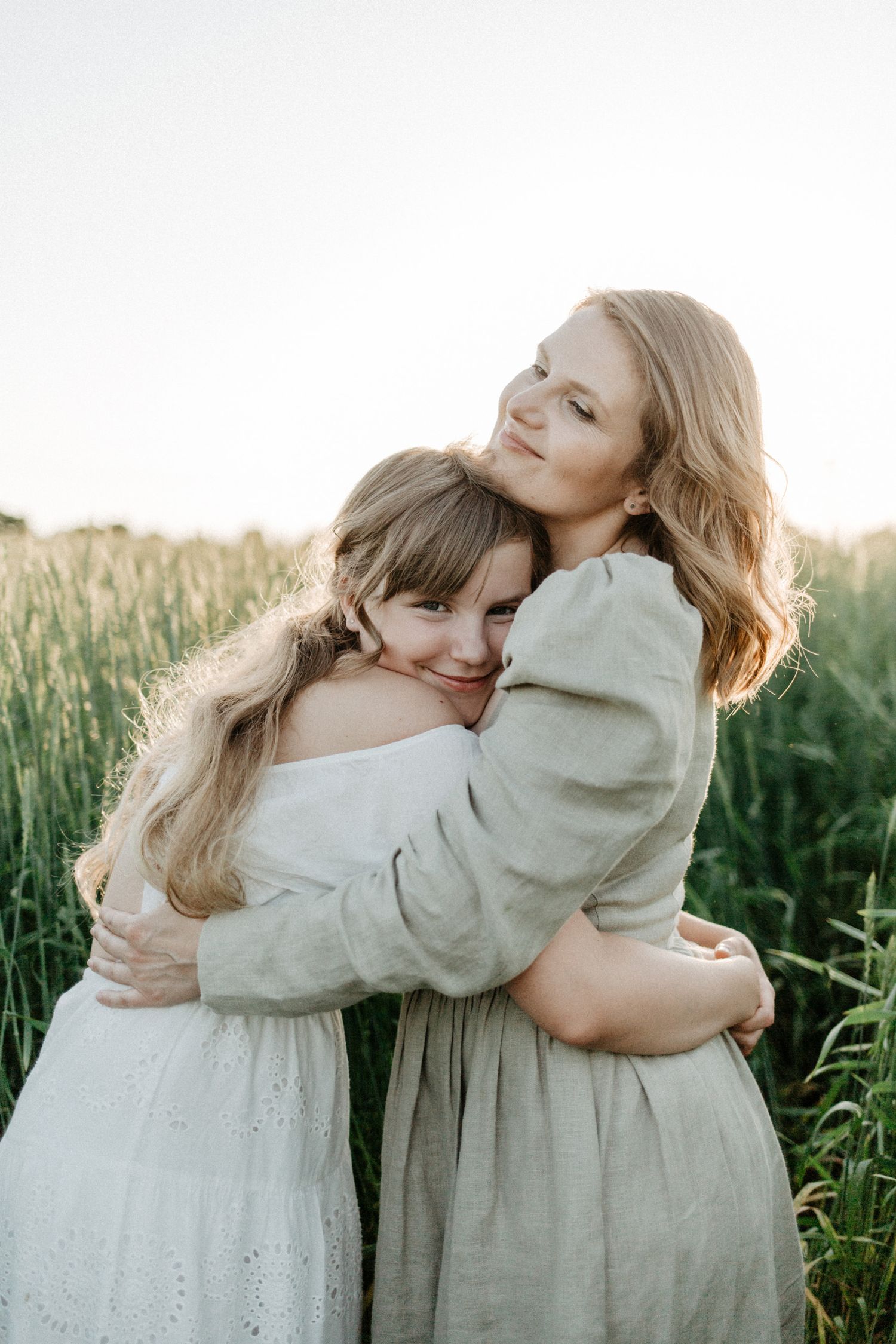 Two people in light colored dresses embracing each other in a sunlit field during golden hour.