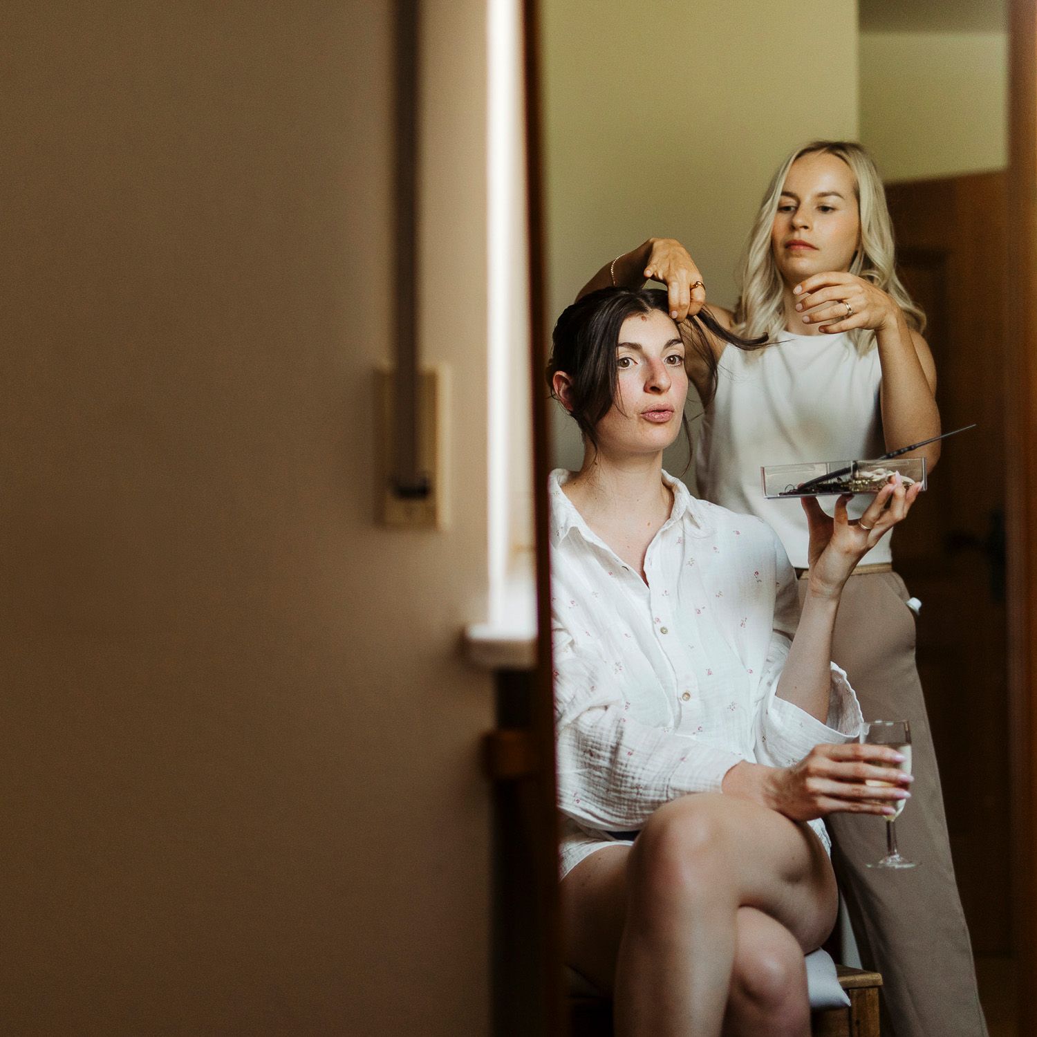Person in white shirt standing by window in moody lighting holds a beverage while looking down contemplatively.