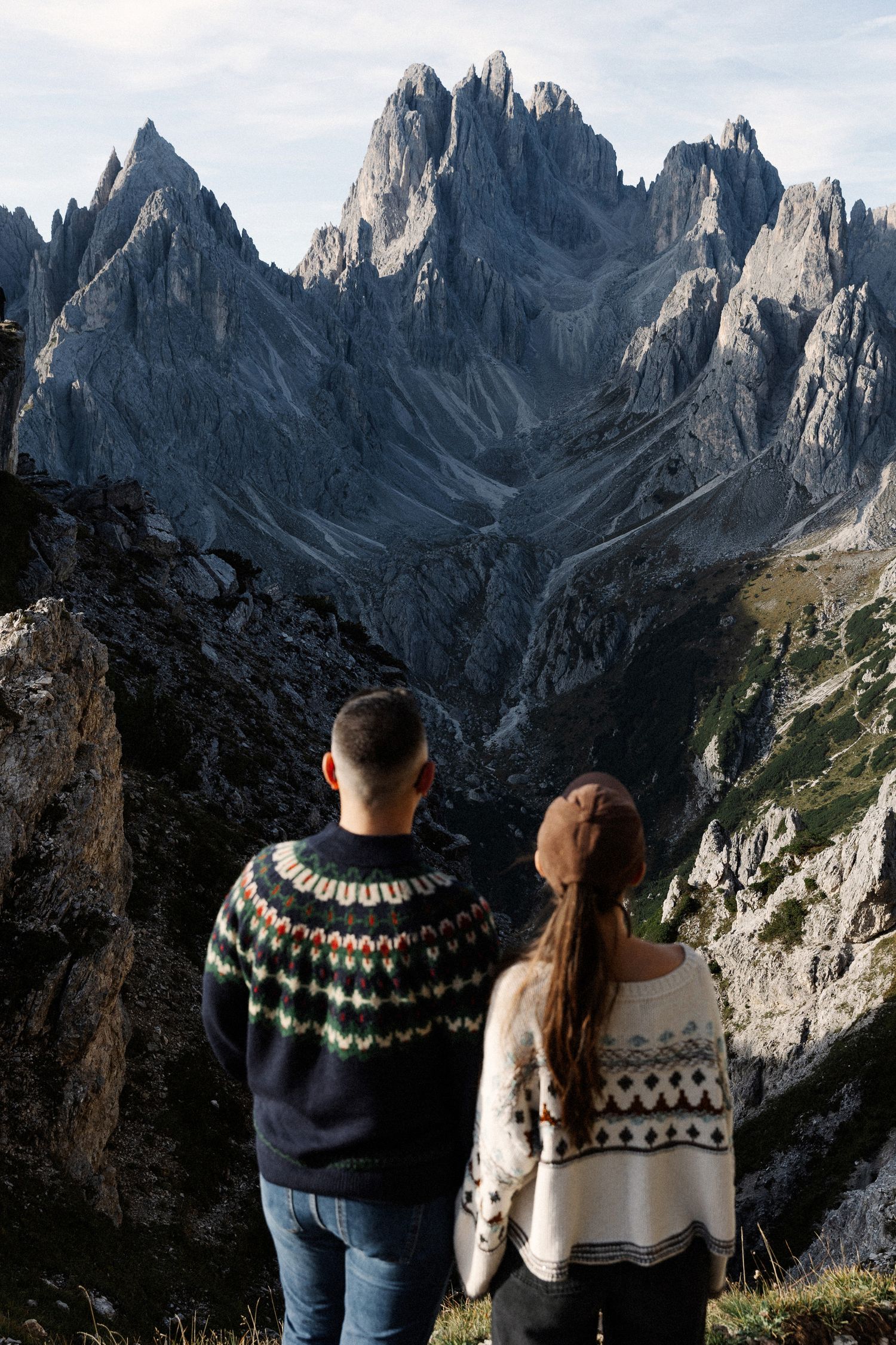Couple in Nordic sweaters admiring dramatic rocky Dolomite mountain peaks from a scenic overlook.