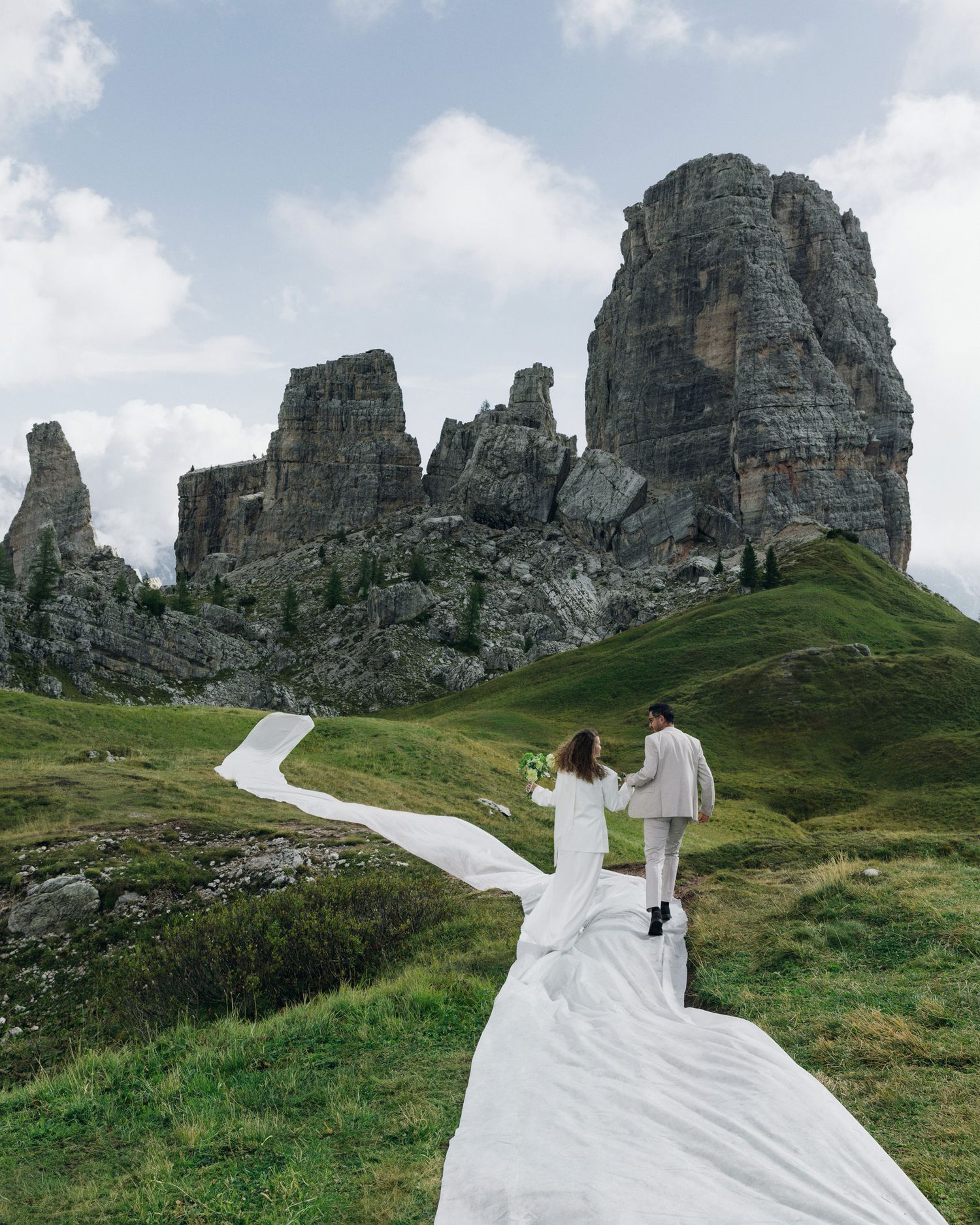 Bride and groom walk hand-in-hand across green alpine meadow with dramatic Dolomite rock formations backdrop.