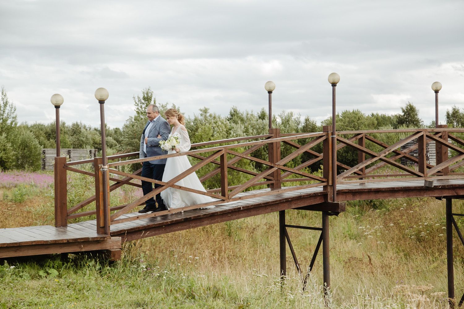 Wedding couple walking across wooden bridge over grassy field.