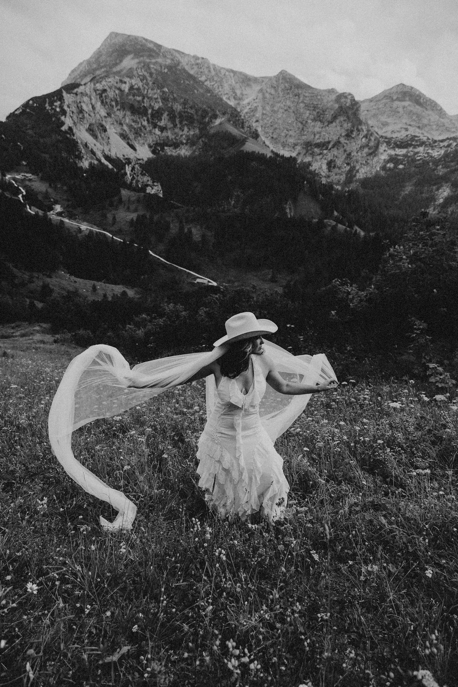 A person in a flowing white dress dances gracefully in a mountain meadow filled with wildflowers, captured in black and white.