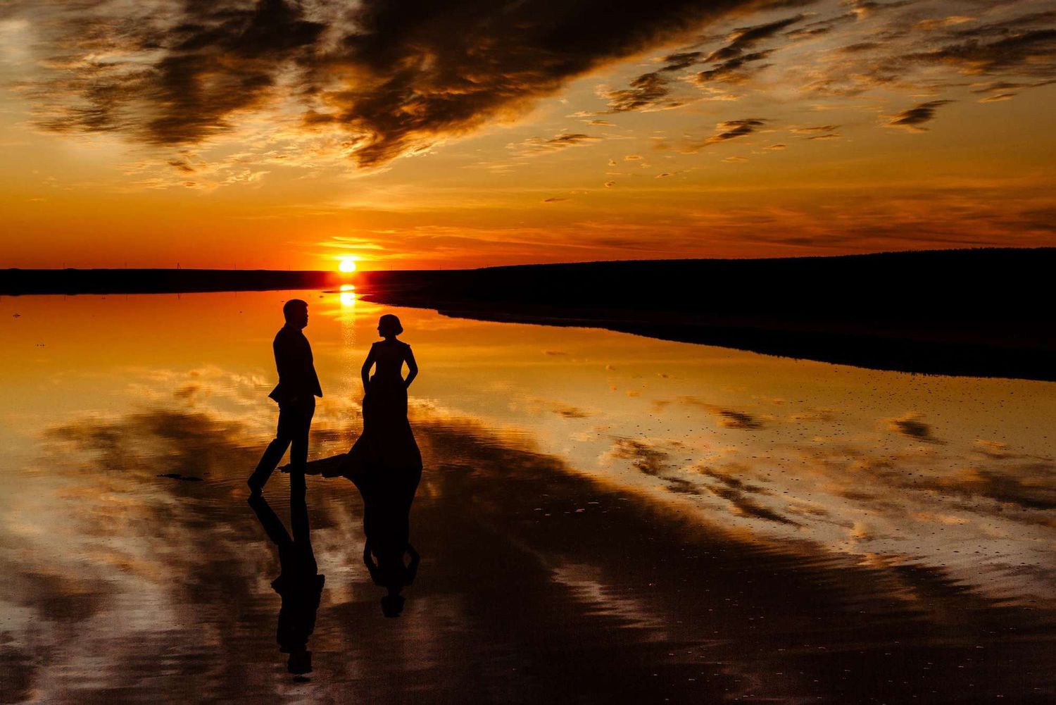 Silhouettes of people walking along a beach at sunset with dramatic orange and golden clouds in the sky.