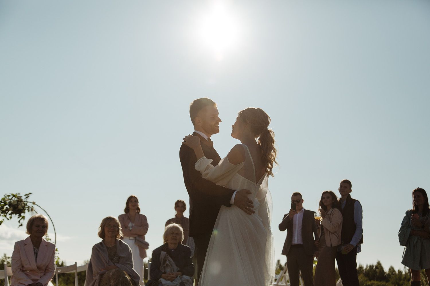 Wedding celebration silhouettes against bright sky during outdoor reception.