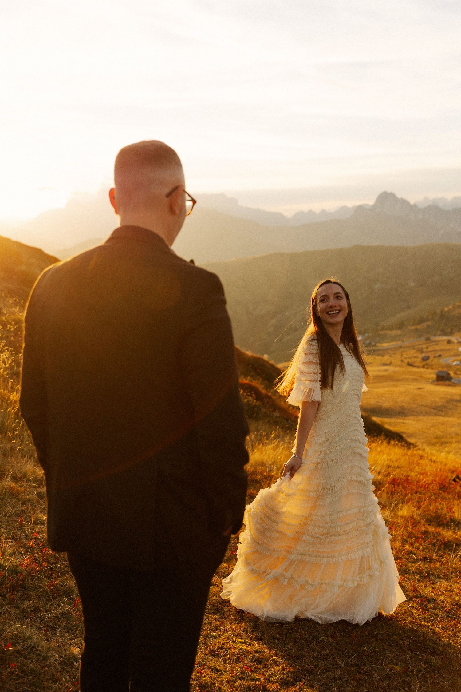 Bride in white dress laughs with groom at golden sunset in mountain meadow during outdoor wedding photo shoot.