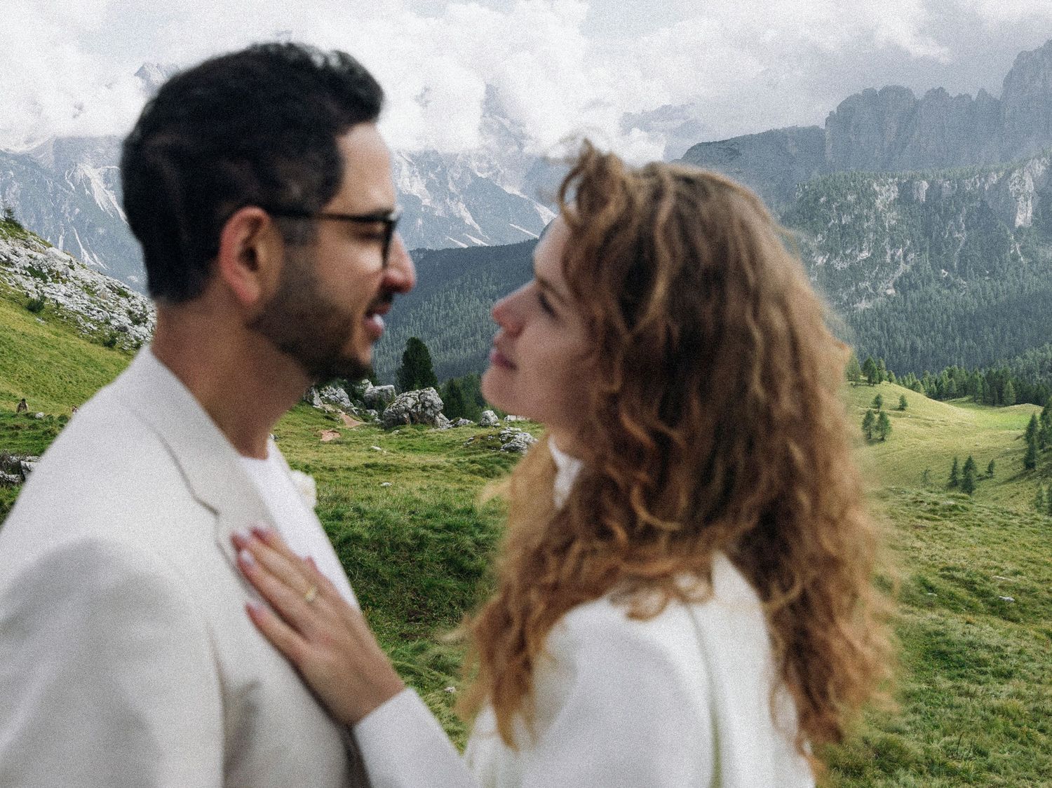 Couple in white attire smiling at each other with scenic Dolomites mountain landscape in background.