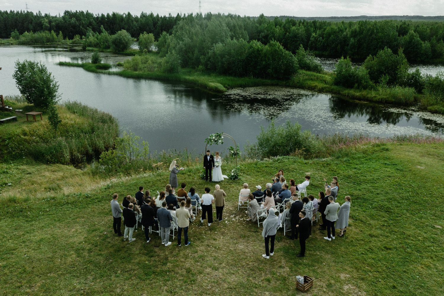 Aerial view of wedding guests gathered near a serene waterfront setting and lush greenery.