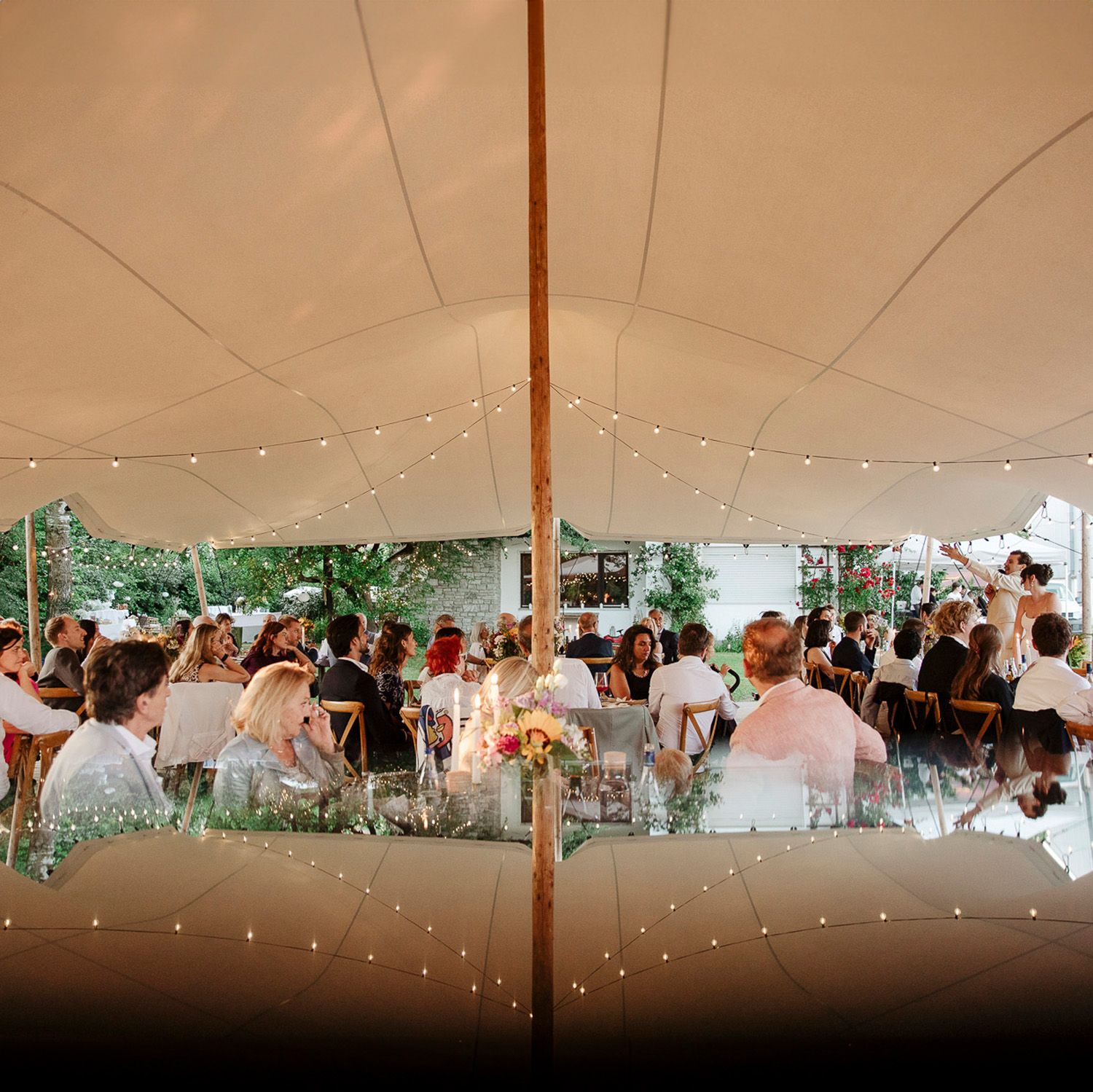 A large white wedding tent filled with guests and decorated with string lights for an outdoor reception.