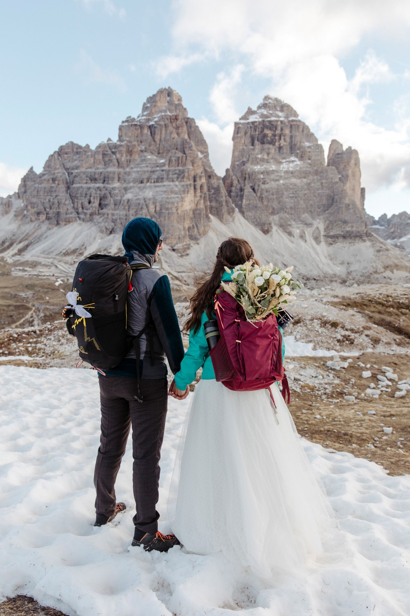 A couple in wedding attire explores a snowy mountain landscape with dramatic Dolomite peaks in the background.