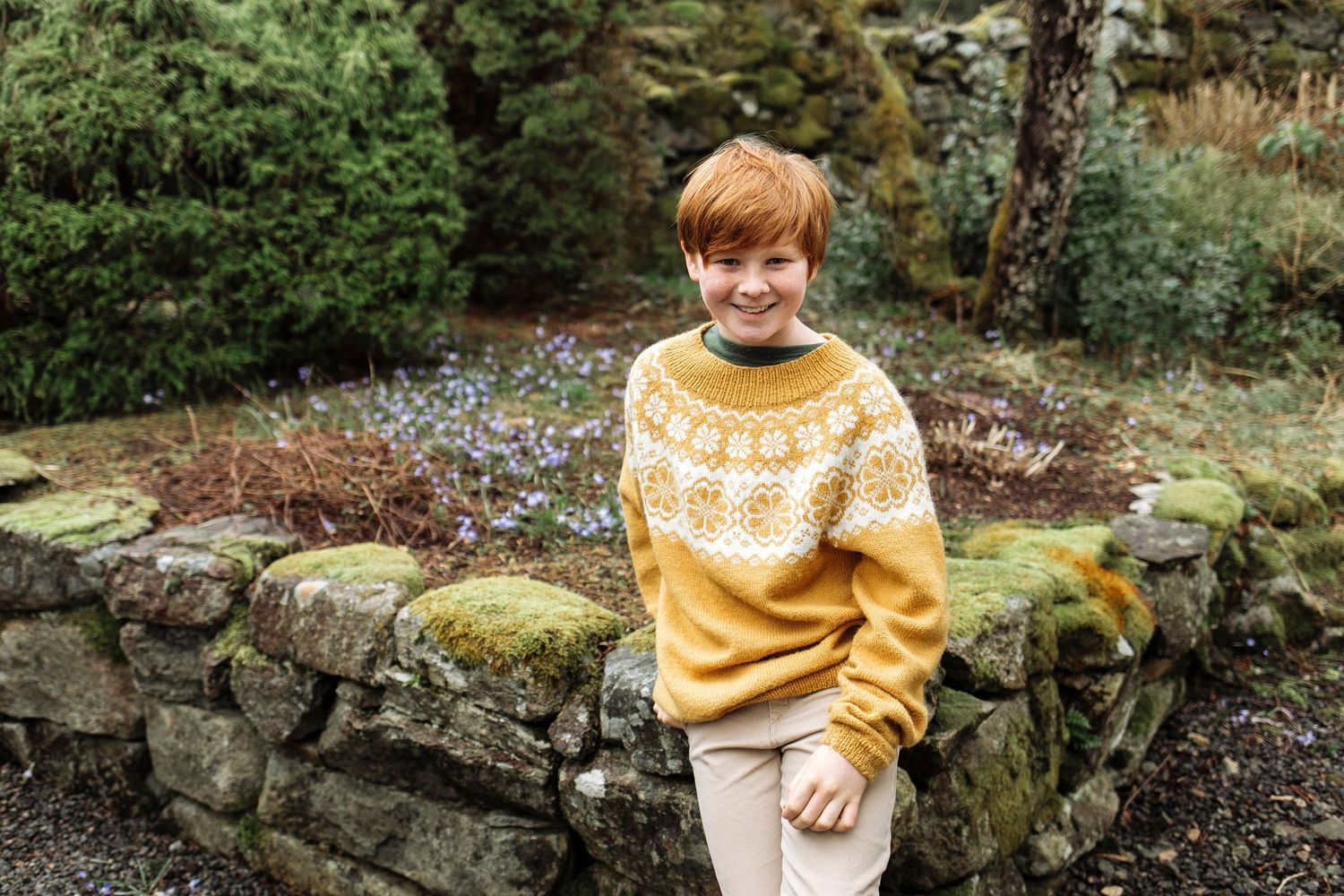 Cozy fair isle pattern yellow sweater worn against a mossy stone wall backdrop in a garden setting.