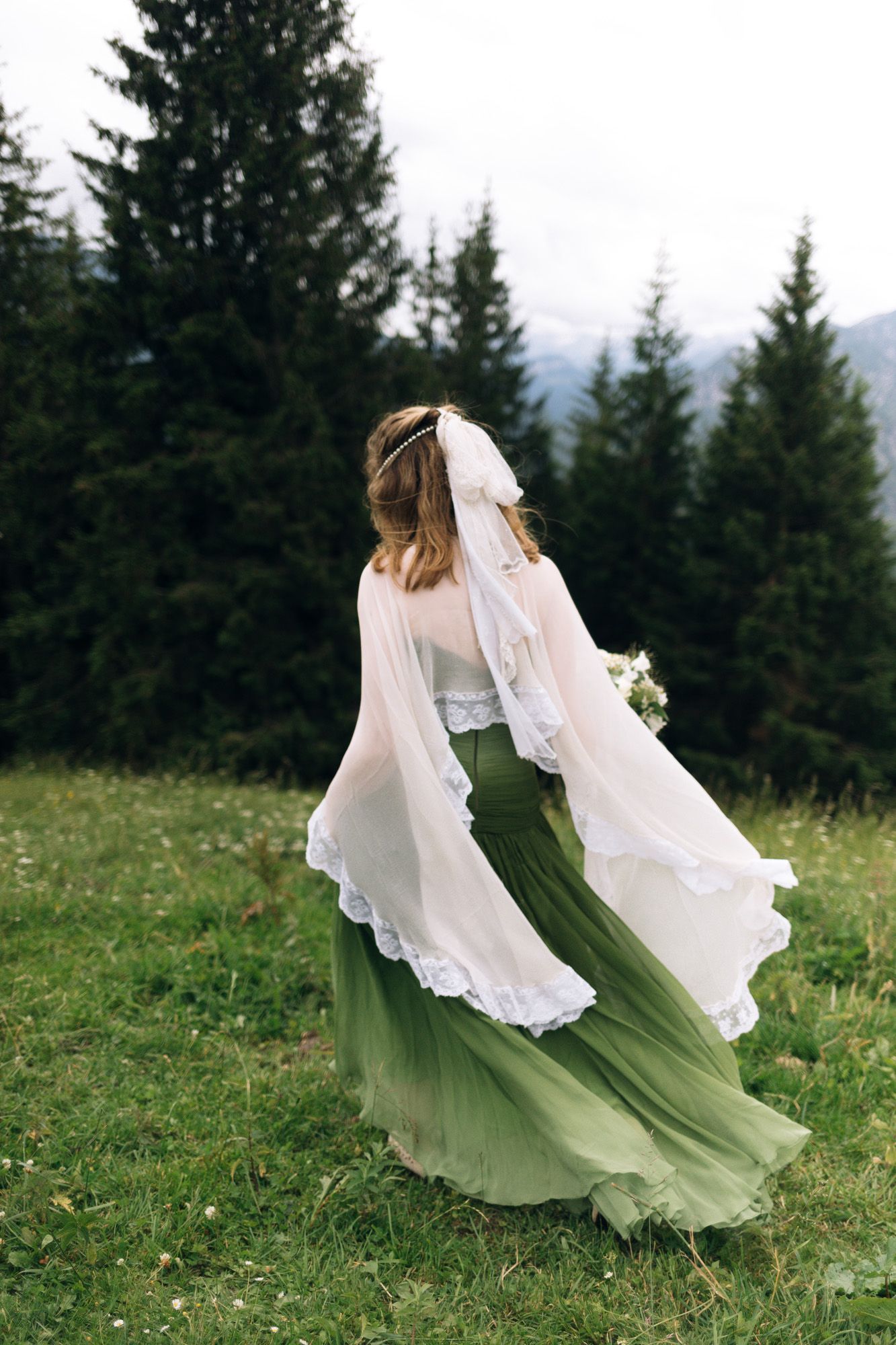 A flowing green and white wedding dress billows gracefully in the wind against an evergreen forest backdrop.