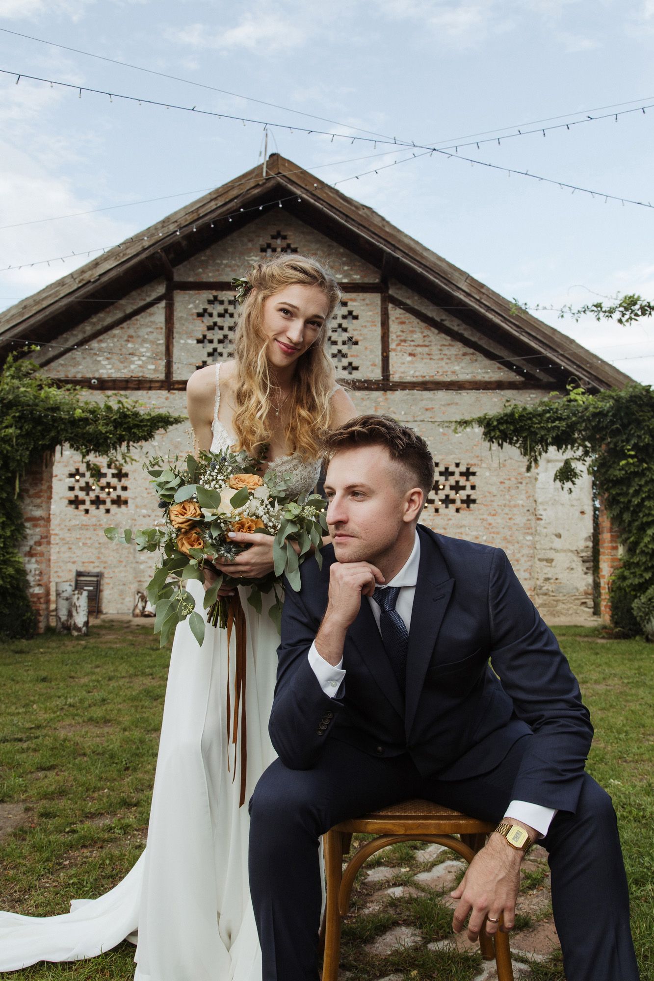 A rustic wedding portrait in front of an old stone building with string lights overhead.