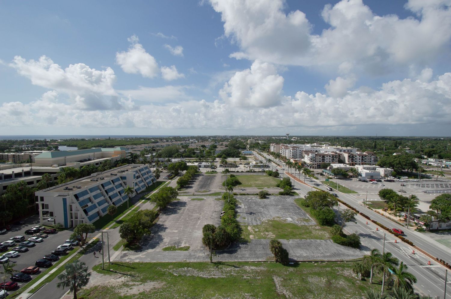 Aerial view of a large vacant lot in a suburban Florida city with scattered trees, parking areas, and buildings under cloudy skies.
