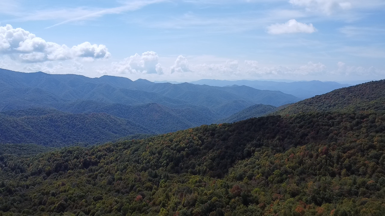 Panoramic view of lush green Appalachian mountain ridges under a partly cloudy blue sky with autumn foliage.