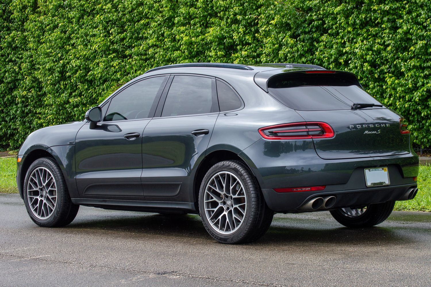 Dark gray Porsche Macan SUV rear three-quarter view parked on wet pavement with green hedge background.