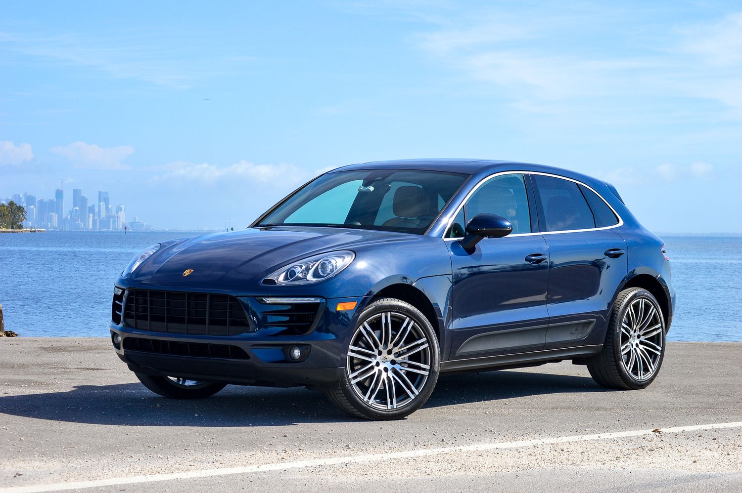 Dark blue Porsche Macan SUV parked near waterfront with city skyline in background on a clear day.