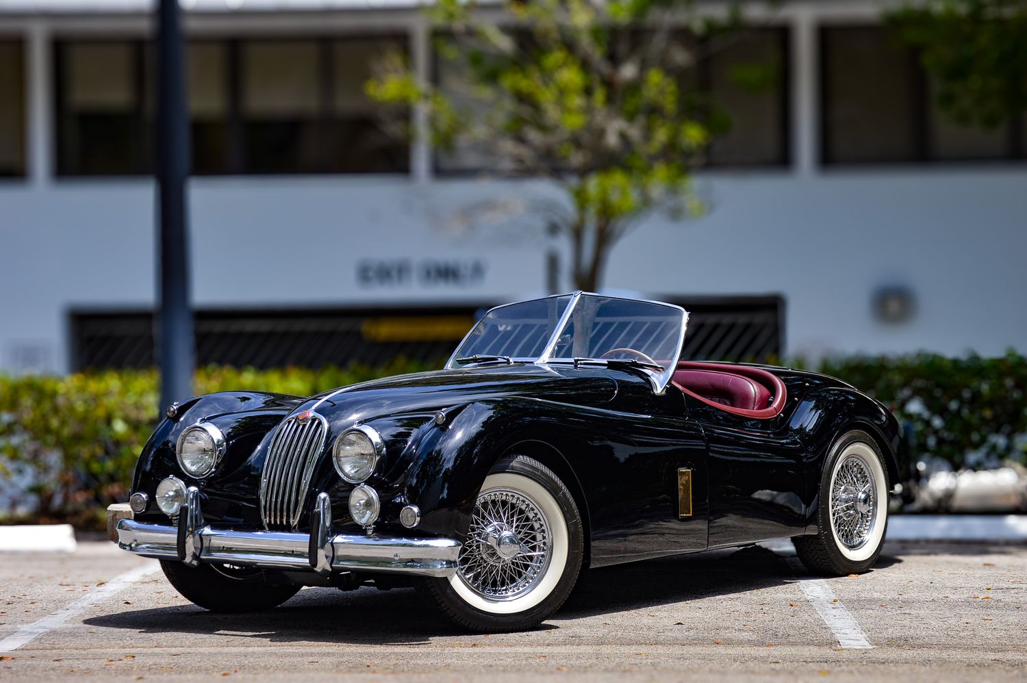 Classic black Jaguar XK140 roadster convertible with wire wheels and whitewall tires parked in a lot.
