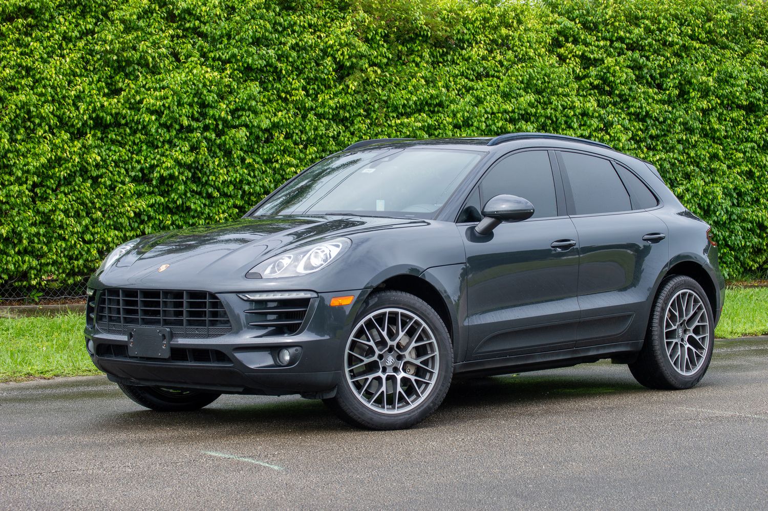 Dark gray Porsche Macan SUV parked on pavement with lush green hedges in the background.