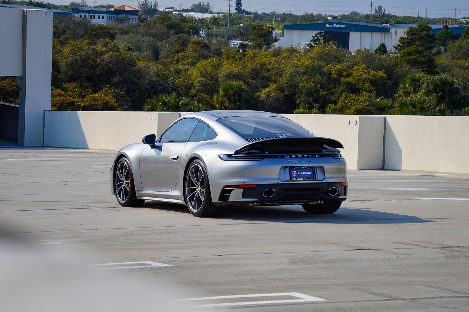 Silver Porsche 911 Turbo S parked on rooftop parking deck with trees and blue sky in background.