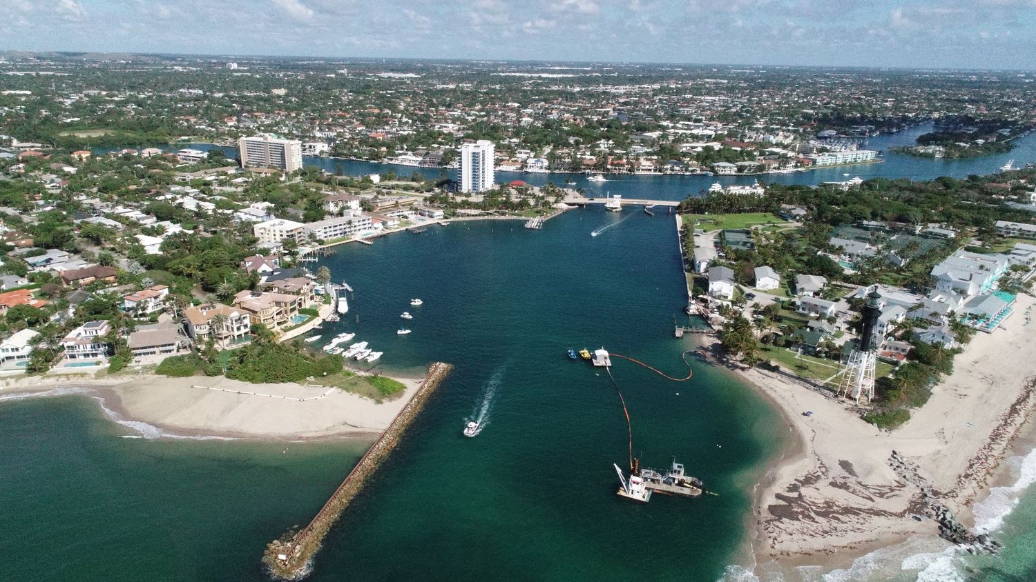 Aerial view of a coastal Florida city with turquoise harbor waters, boats, sandy shores, and urban skyline on a sunny day.