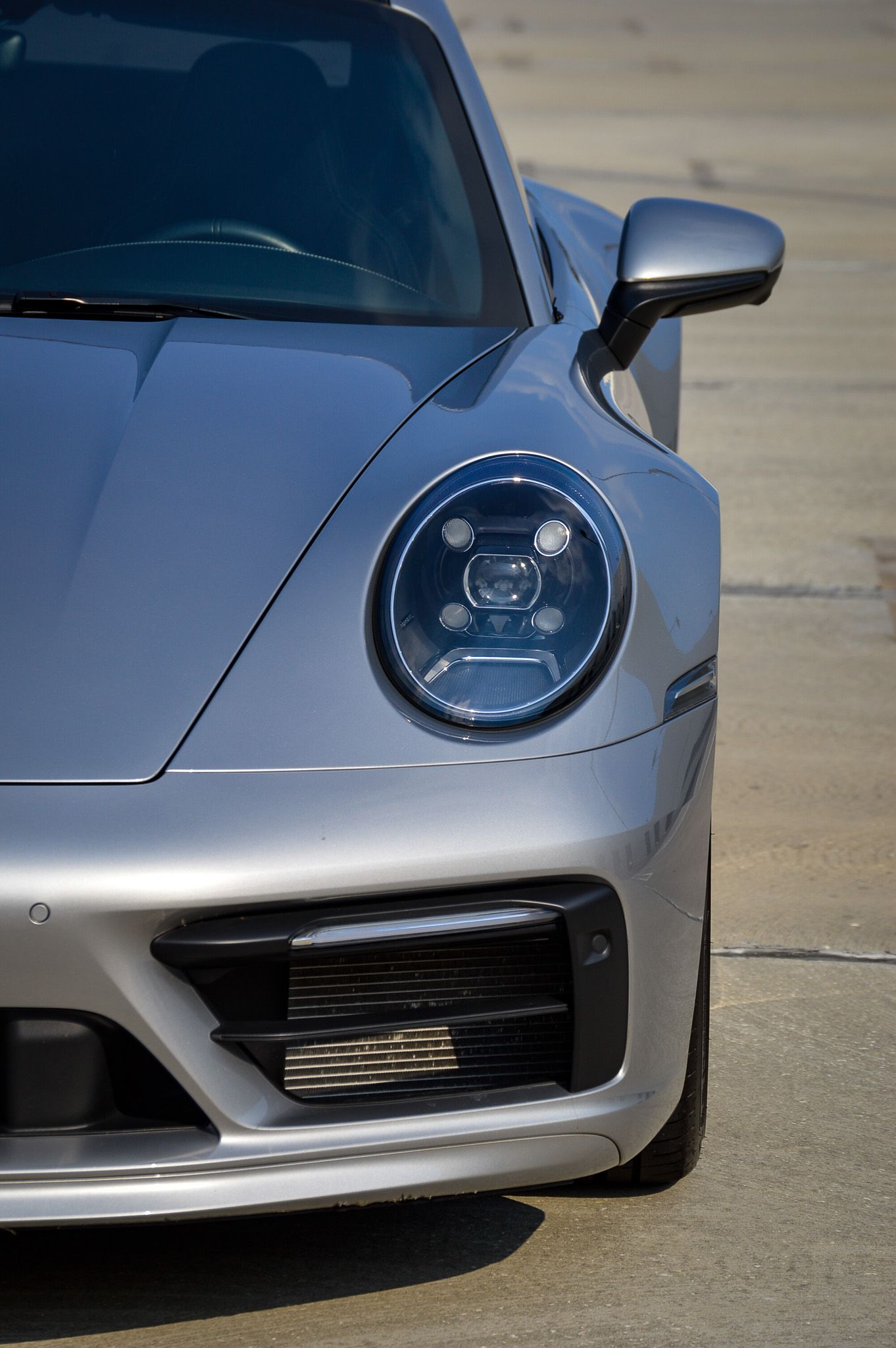Silver Porsche 911 front quarter view showcasing LED headlight and sleek aerodynamic bodywork on a concrete surface.