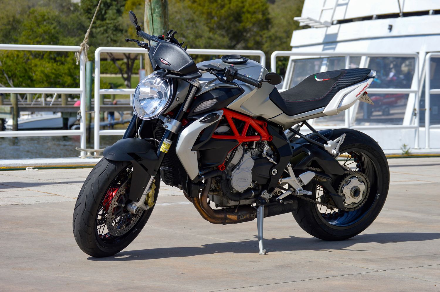 A sleek black and red Ducati Monster motorcycle parked on a waterfront plaza with trees and a boat in the background.