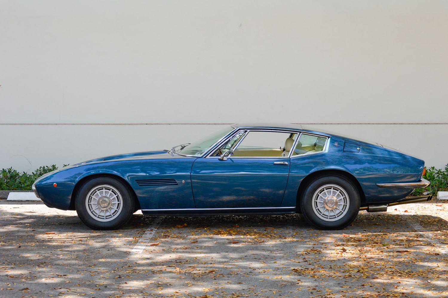 Blue vintage Maserati Ghibli coupe parked beside a white wall, showcasing classic Italian sports car design.
