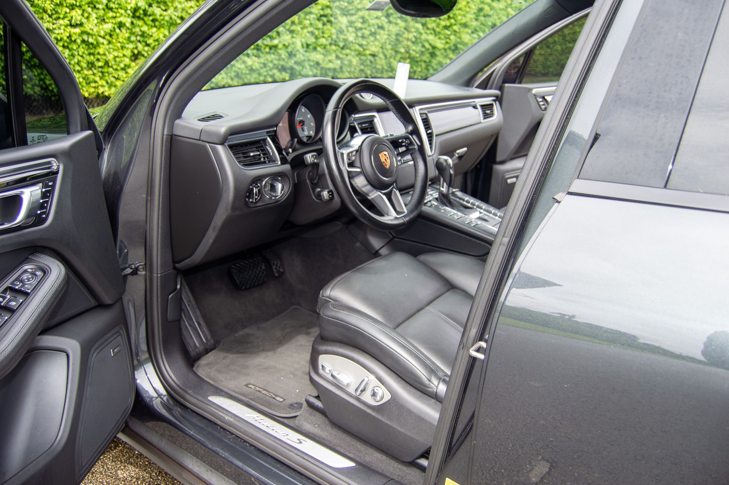 Porsche Macan interior showing black leather seats, sport steering wheel, and premium dashboard with open driver door.