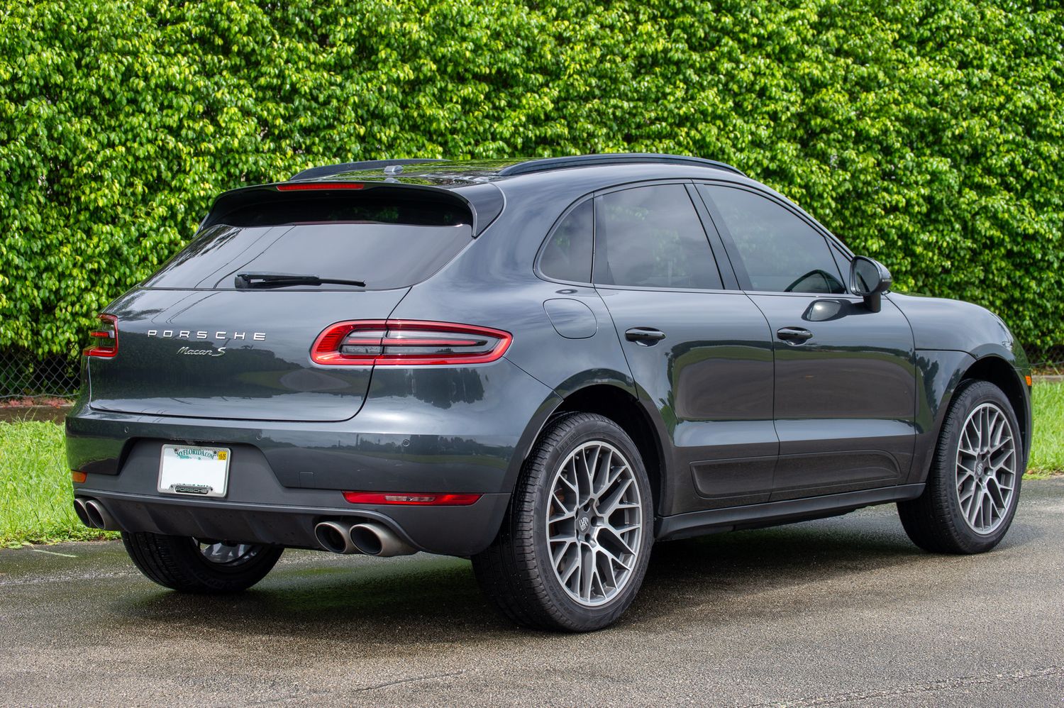 Dark gray Porsche Macan SUV rear view parked on a driveway surrounded by lush green hedges.