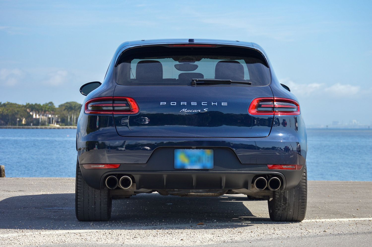 Rear view of a dark blue Porsche Macan S SUV parked near waterfront on a sunny day.