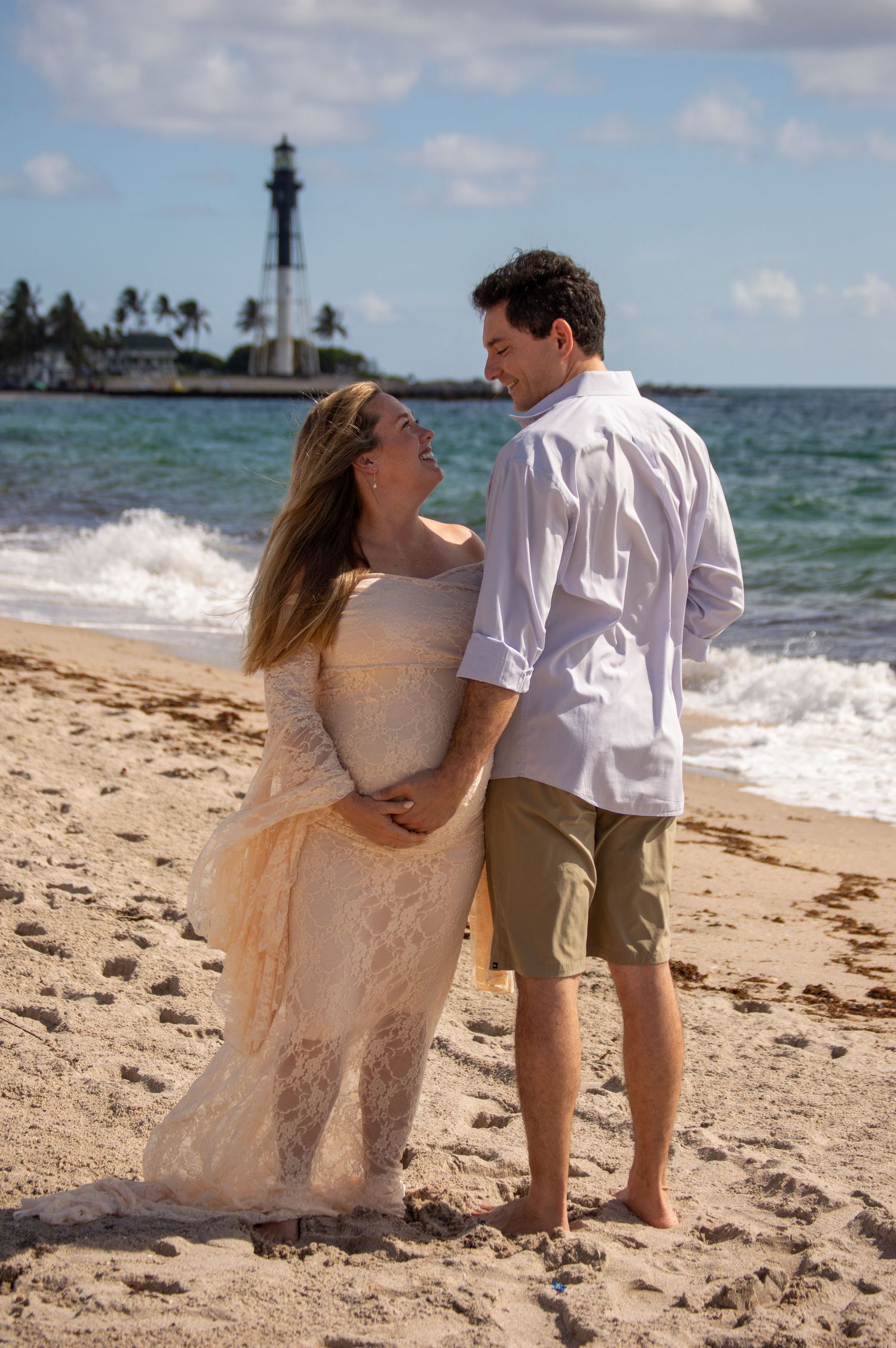 Pregnant couple embracing on sandy beach with Hillsboro Lighthouse in background, woman in lace gown, man in white shirt.