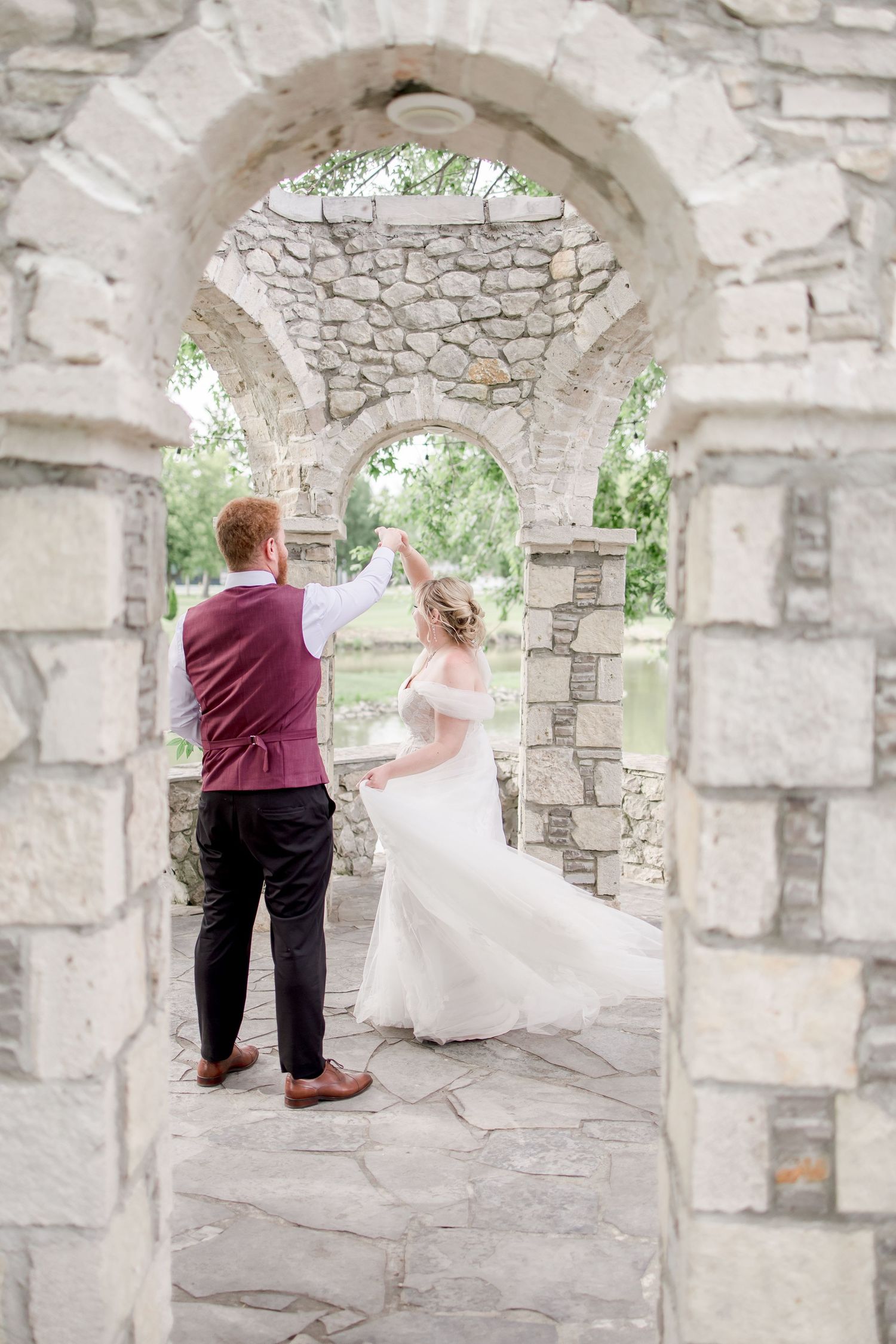 Wedding couple poses beneath rustic stone arches in an outdoor garden setting.