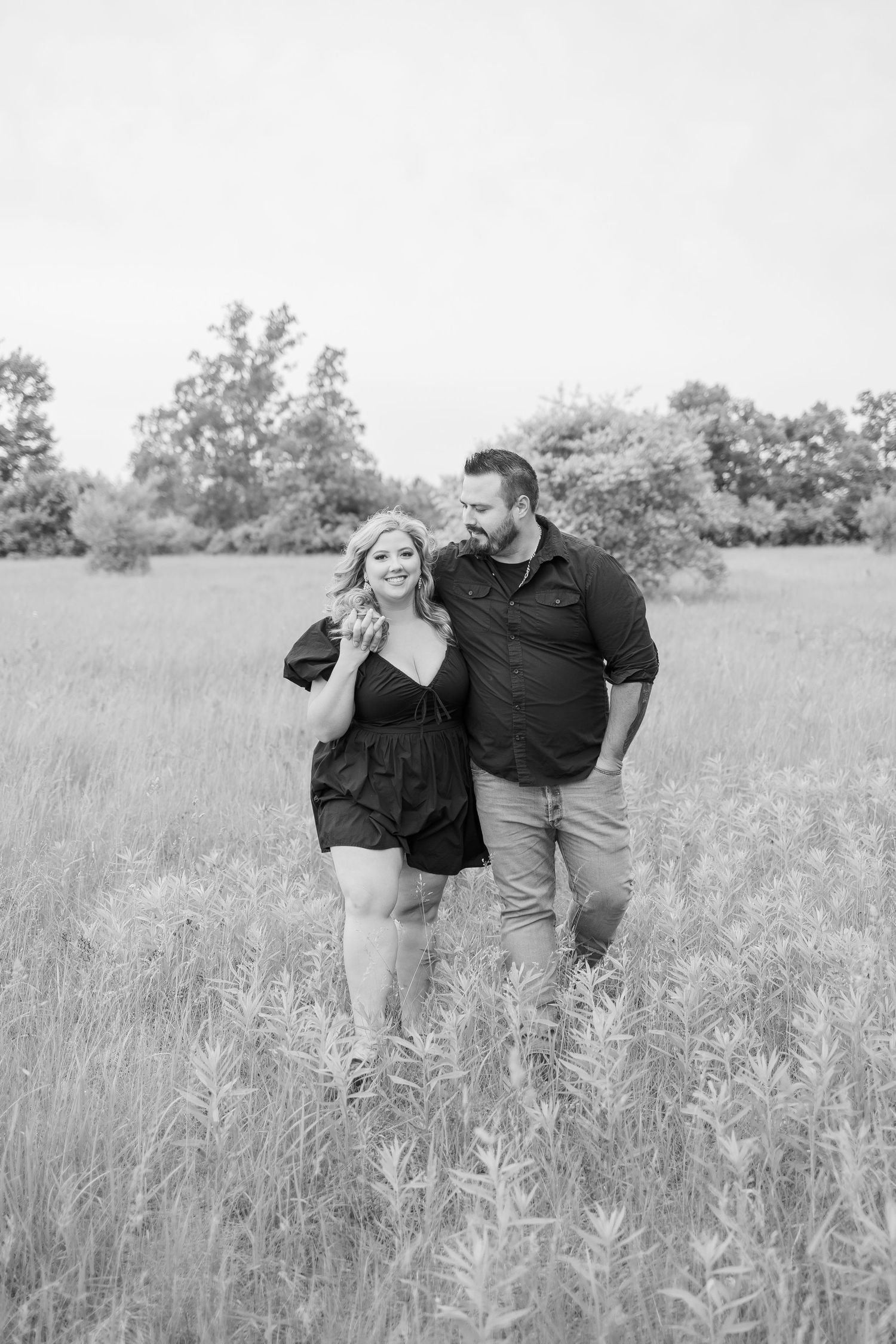 A black and white photo captures a couple's playful interaction in a meadow.