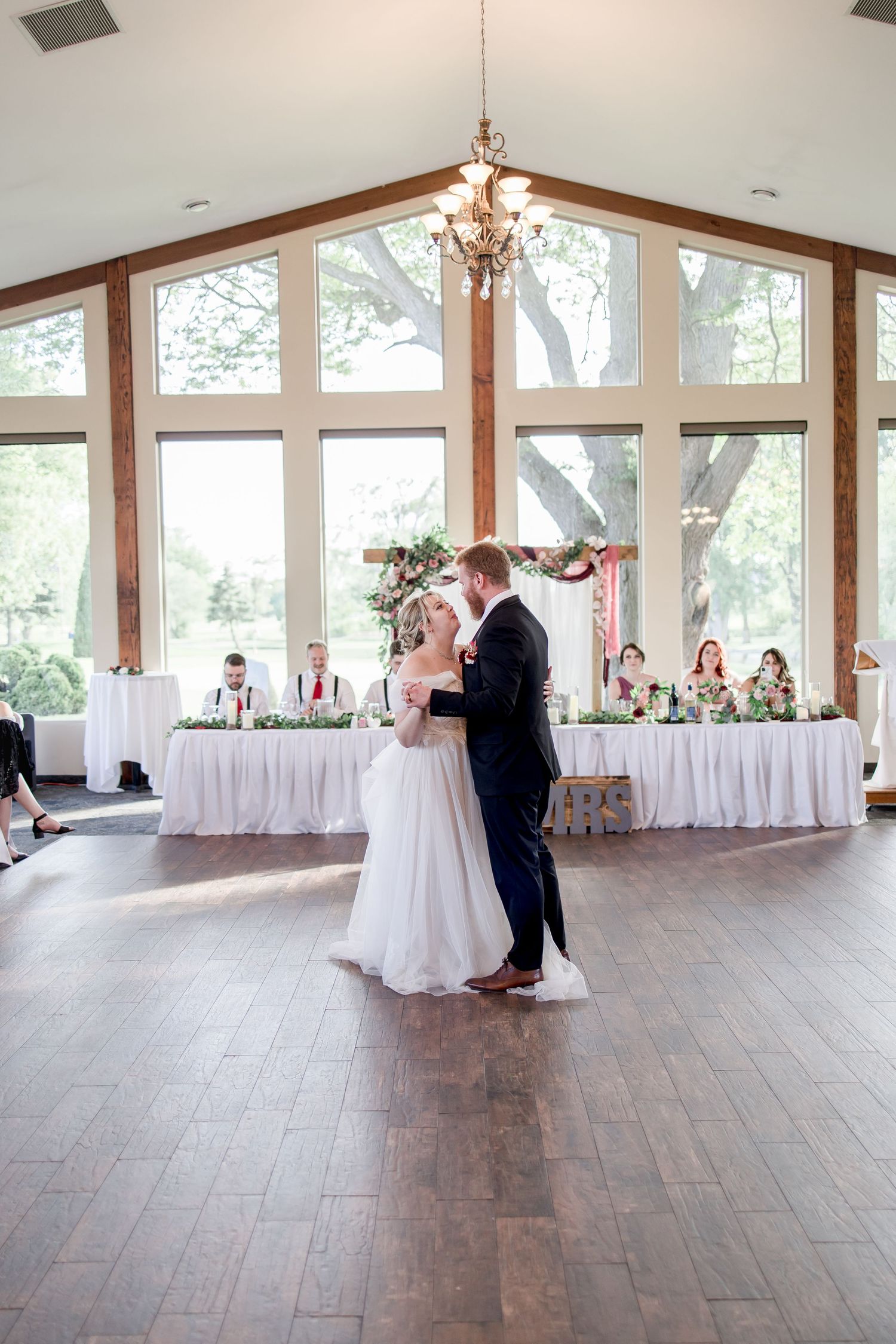 Wedding couple dancing in spacious venue with exposed wooden beams and large windows.