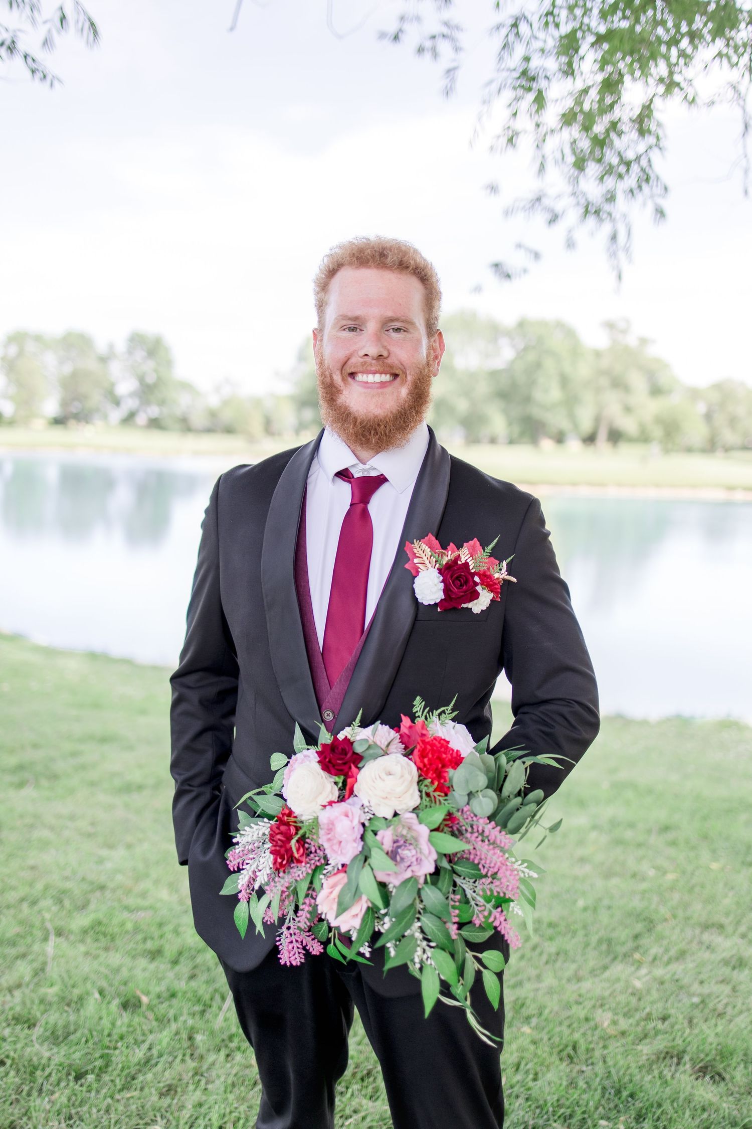 Groom in black suit with red tie holding colorful wedding bouquet.