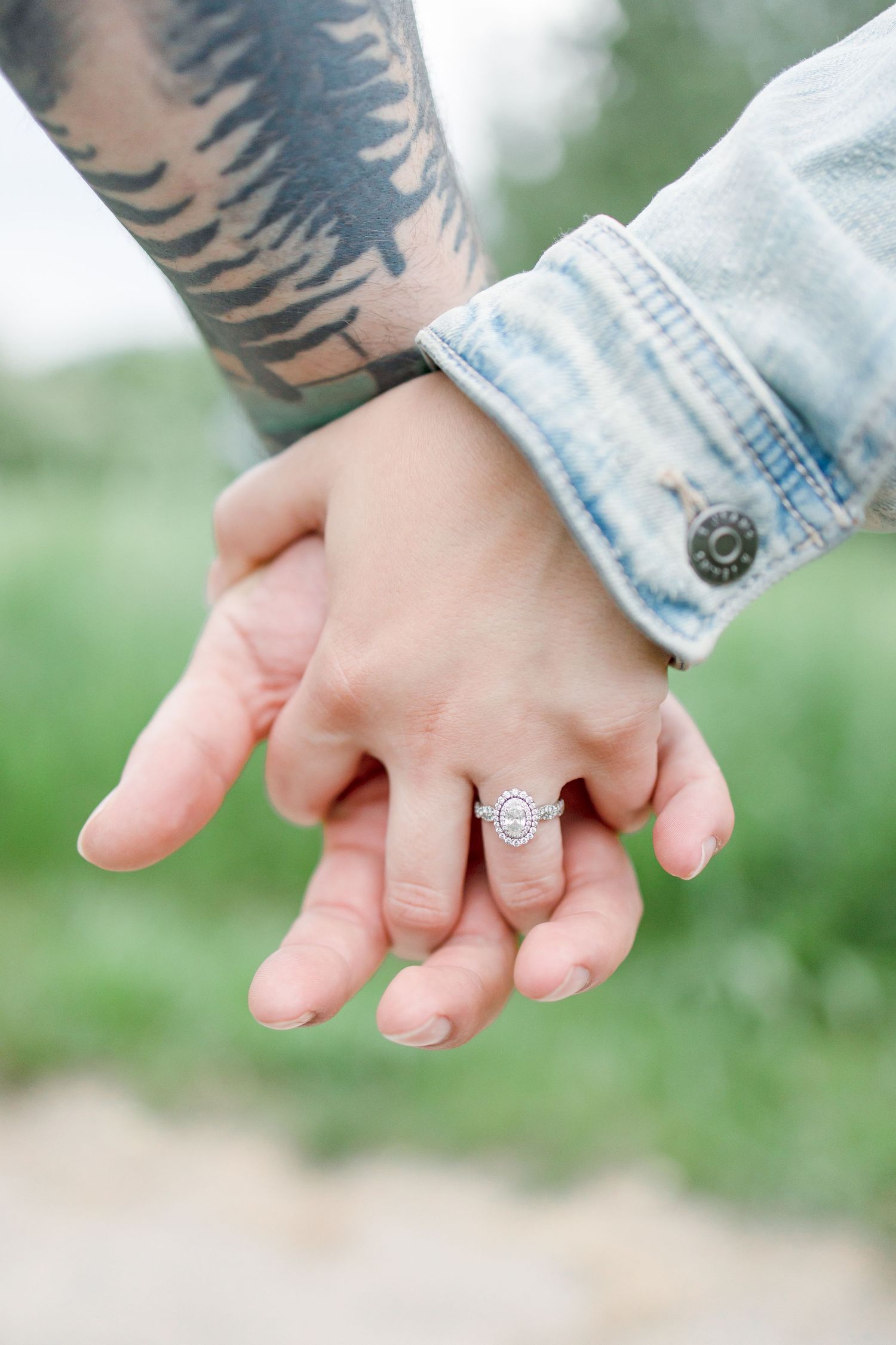 Close-up of clasped hands showing an engagement ring and forest tattoo against denim clothing.