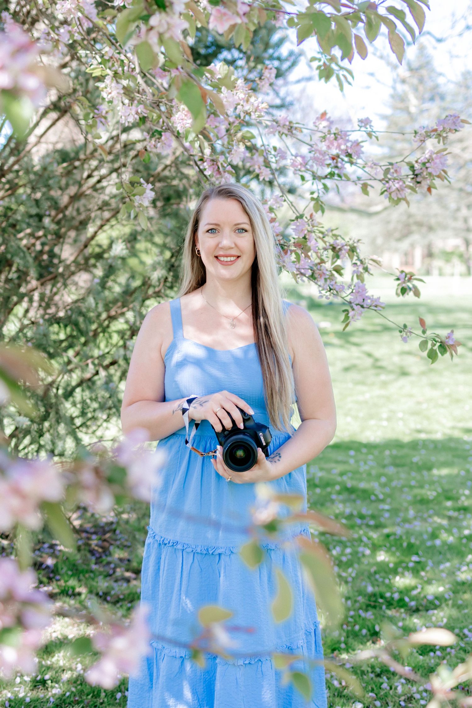 A photographer holds a professional camera while standing under flowering tree branches.