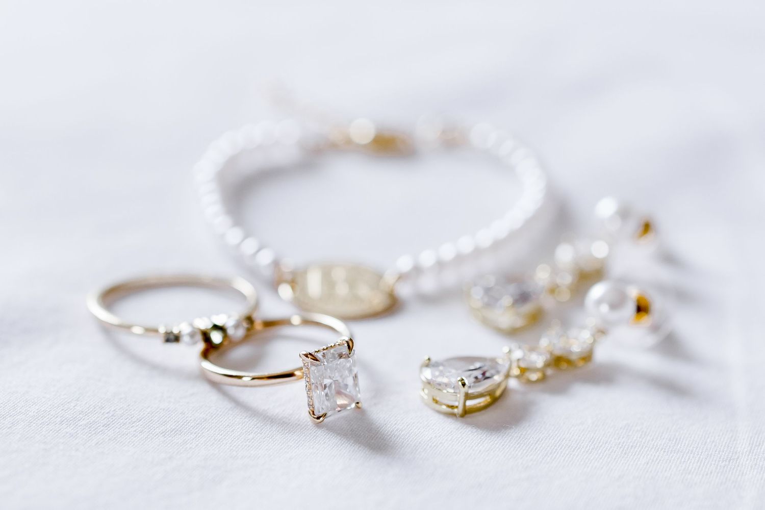 Delicate wedding jewelry including rings and bracelets arranged on white fabric.