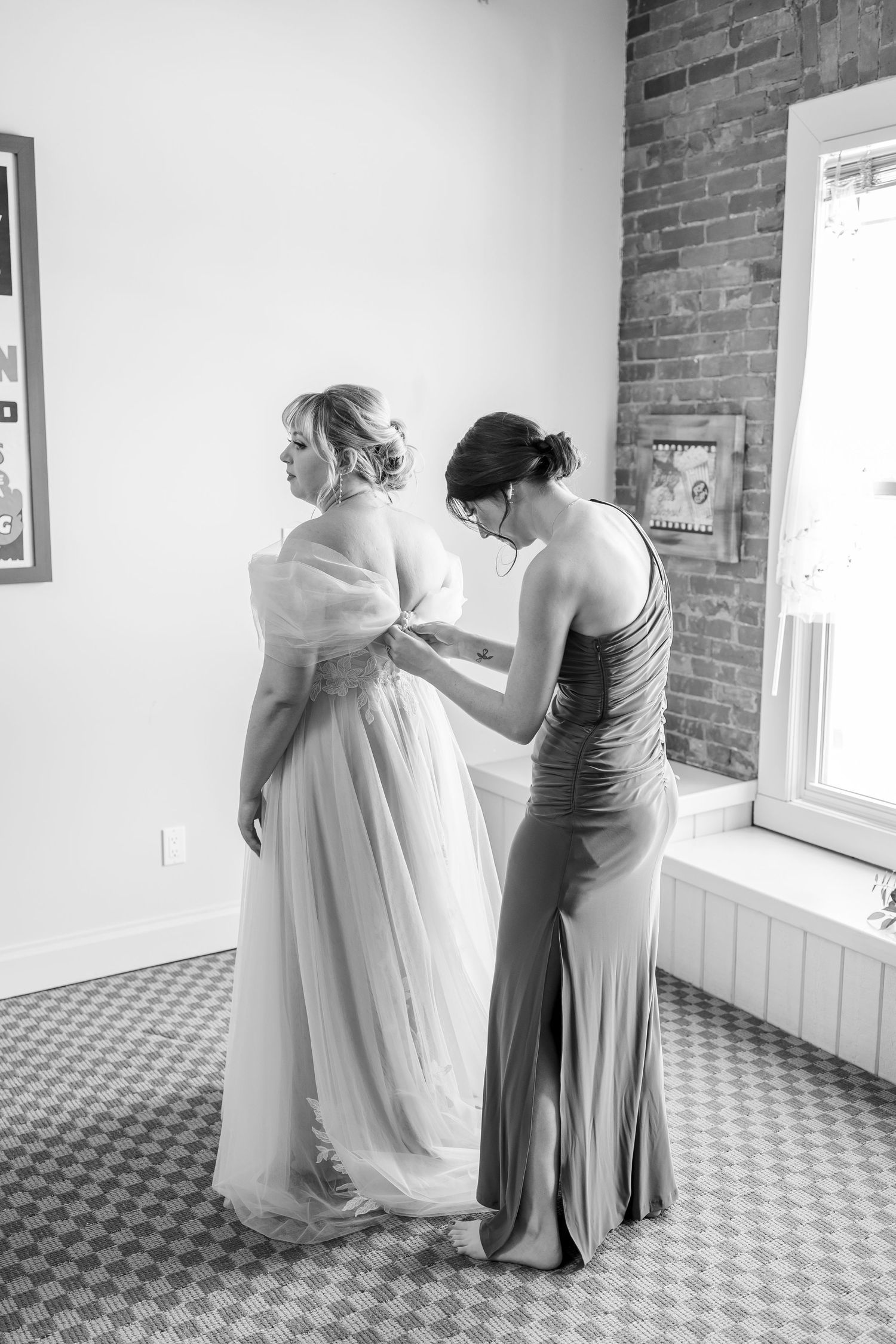 Black and white photo of wedding dress preparation in a room with brick walls and checkered floor.