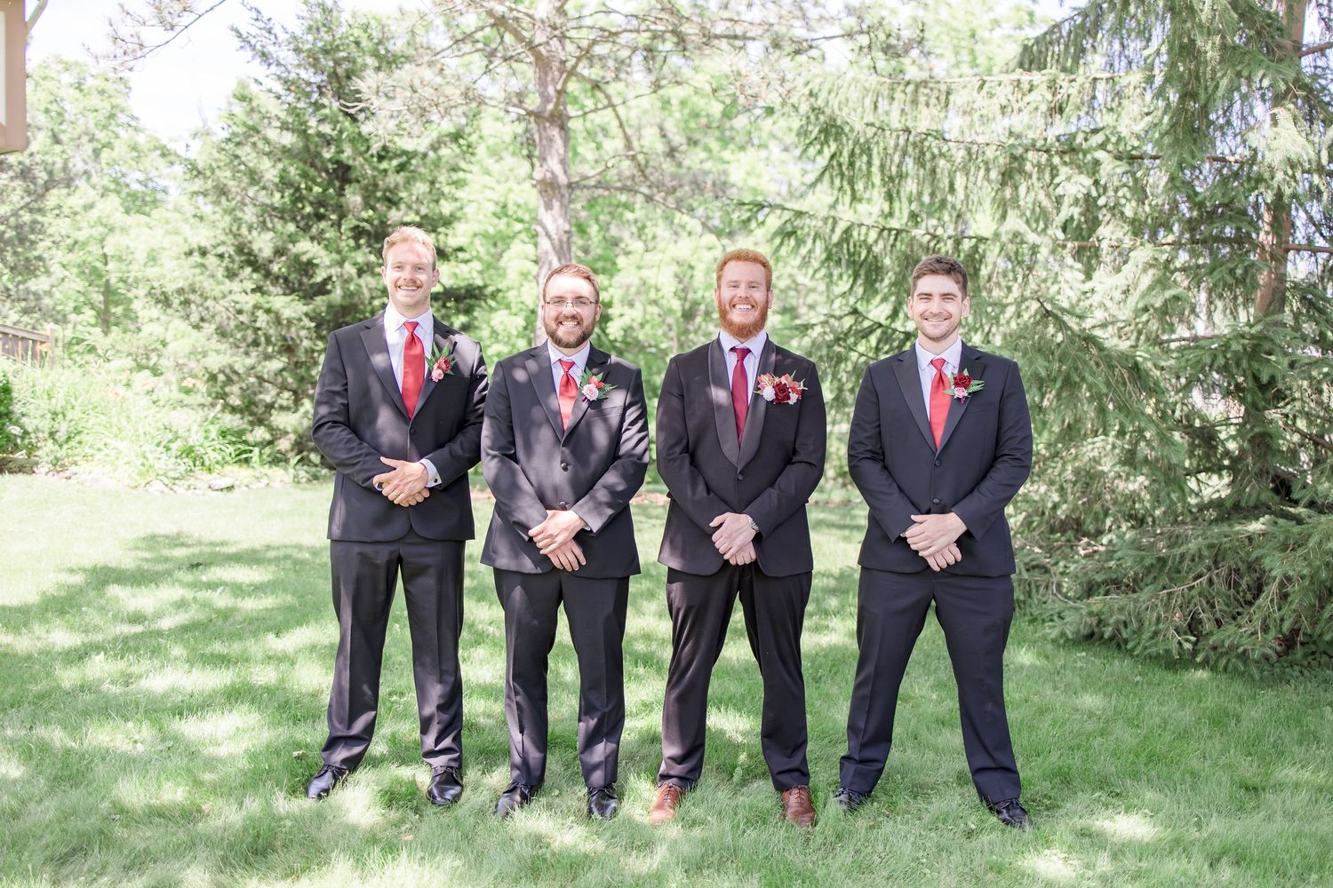 Groomsmen in dark suits and pink ties stand in a line outdoors for formal wedding photos.