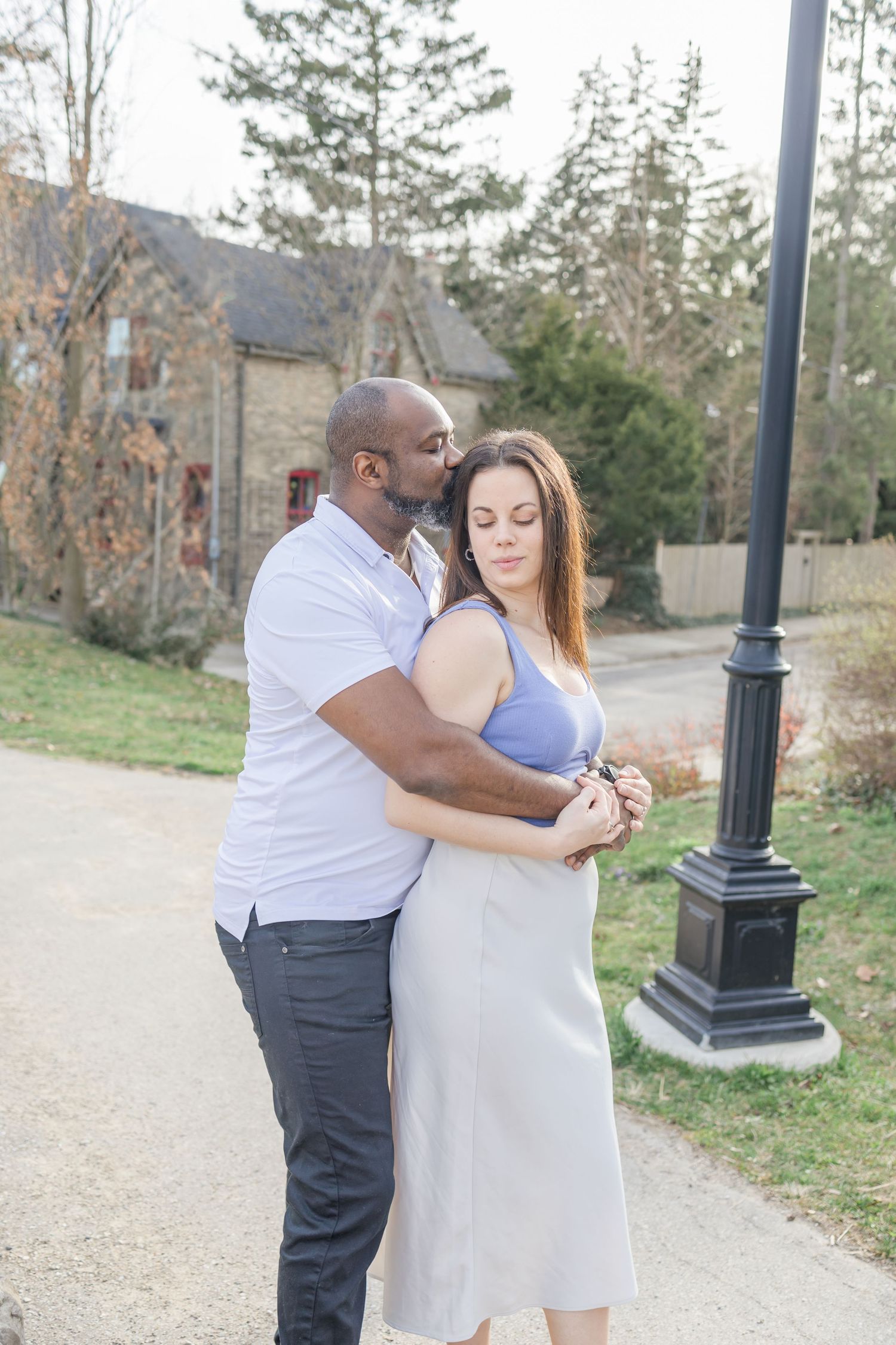Romantic couple embracing in a casual outdoor setting with stone buildings in background.