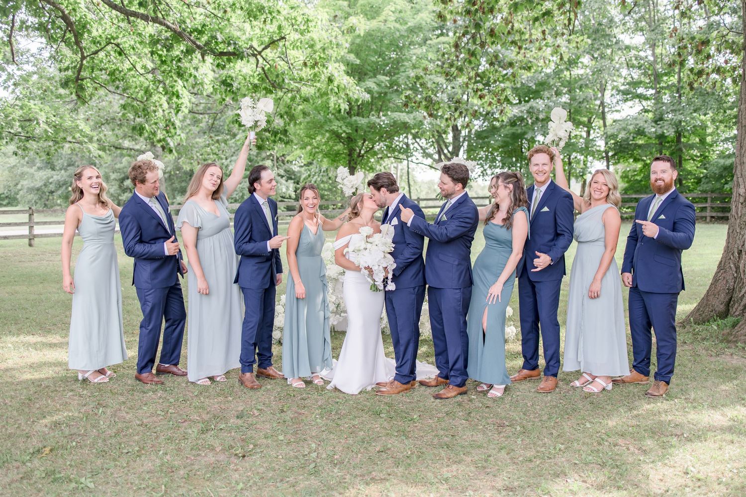 Wedding party in blue suits and light dresses pose together under trees for formal group photos.