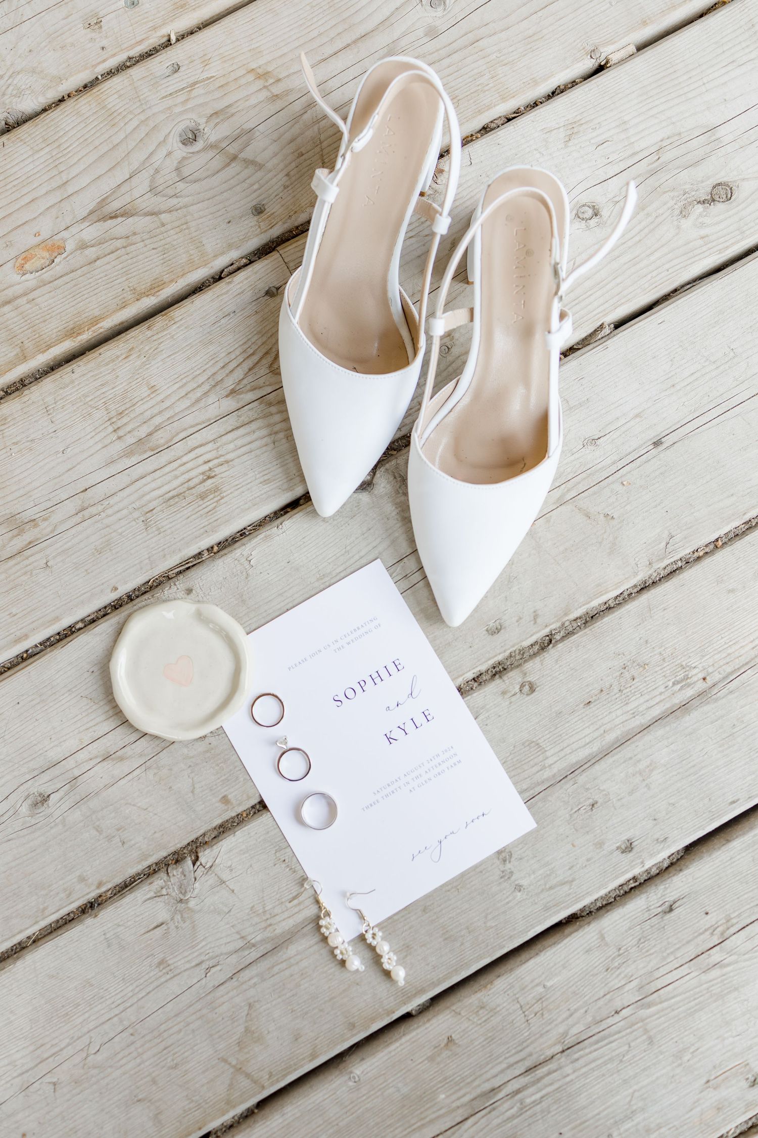 White bridal heels and wedding invitation details arranged on rustic wooden decking.