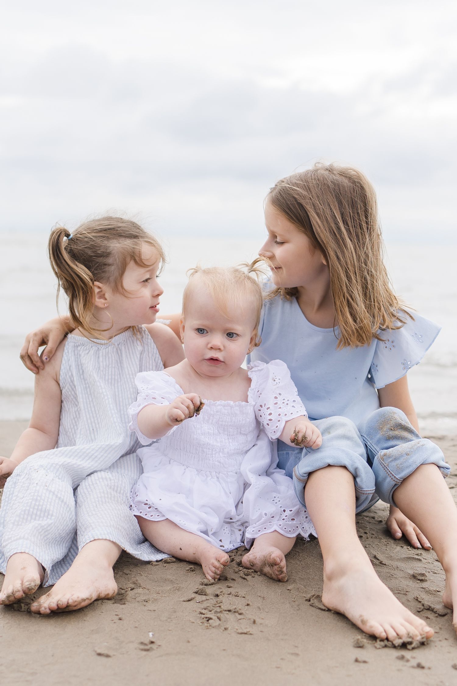 Children sitting together on sandy beach wearing white and blue casual clothing.