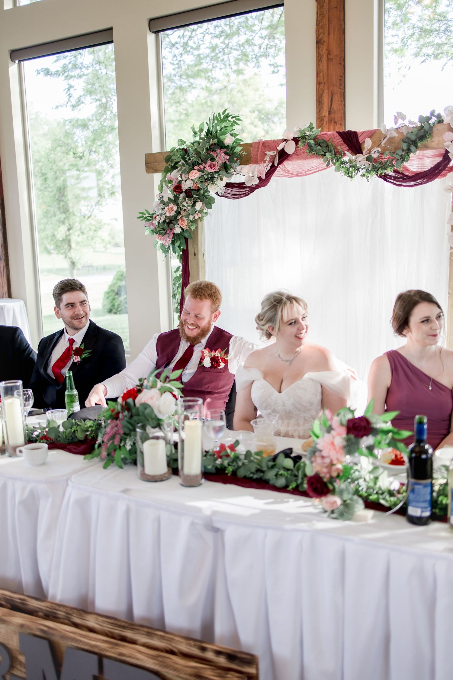 Wedding party seated at decorated head table with burgundy and floral accents during reception celebration.