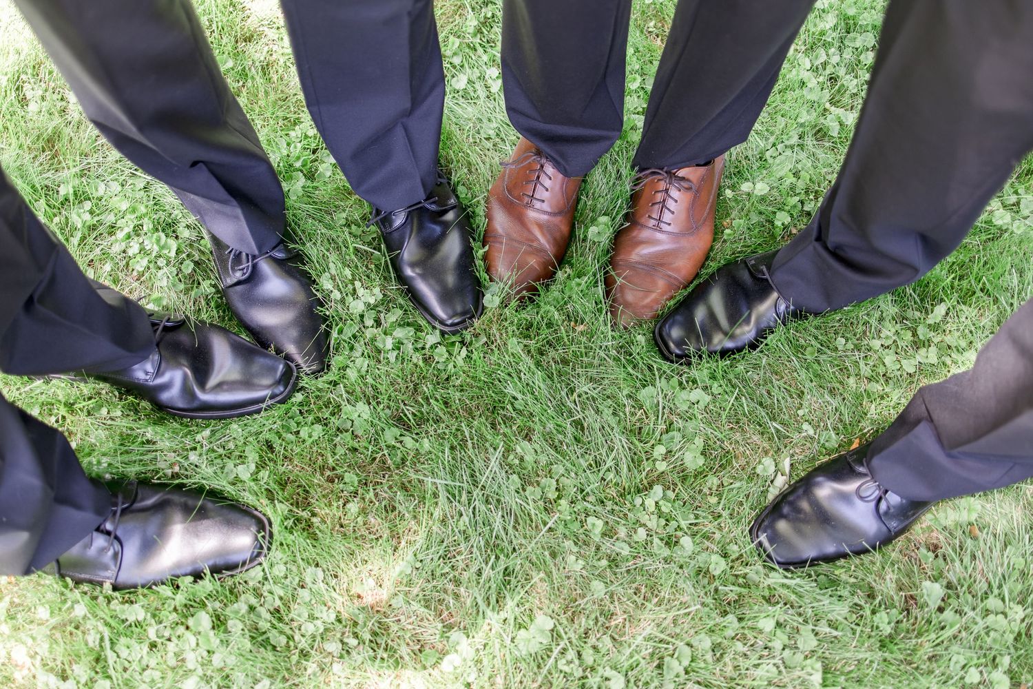 Row of dress shoes and formal footwear lined up on green grass at outdoor wedding.
