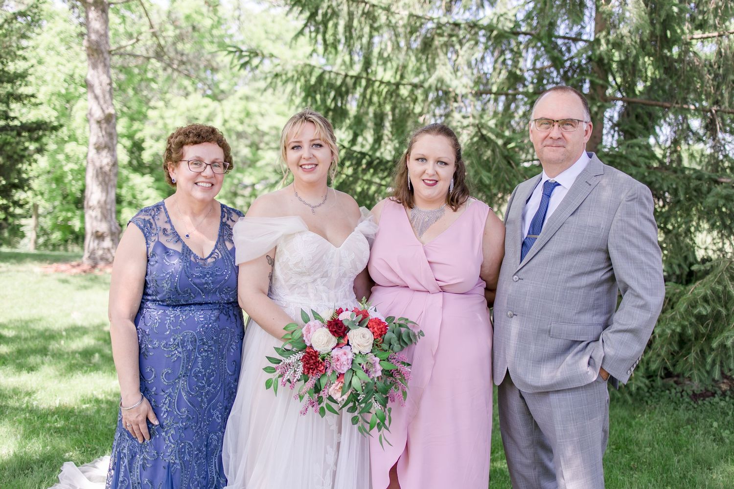 Wedding party members in pink dresses and gray suits pose together outdoors under evergreen trees.