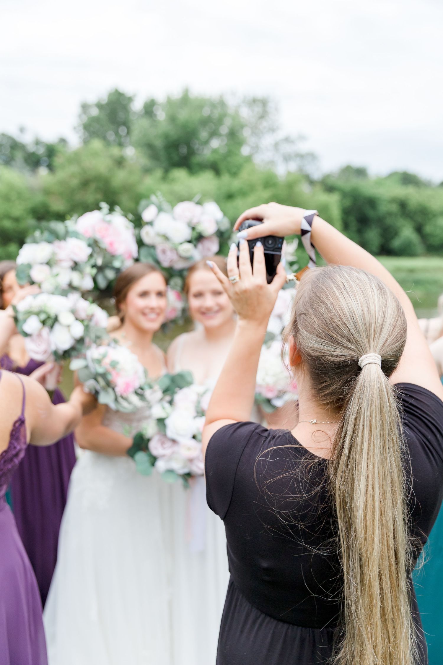 Wedding party members in purple dresses and black attire pose together with floral bouquets outdoors.